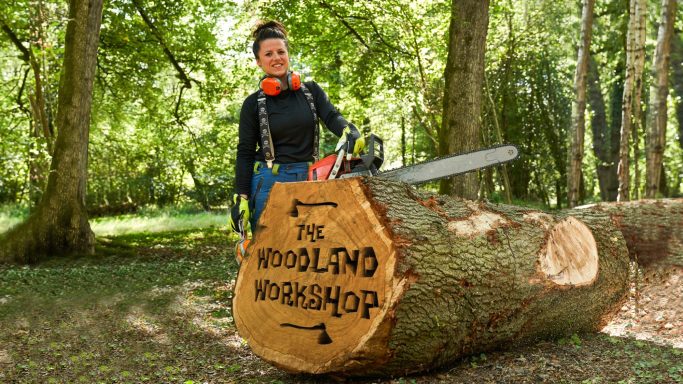 Person with a chainsaw stands beside a large, engraved log in a wooded area.