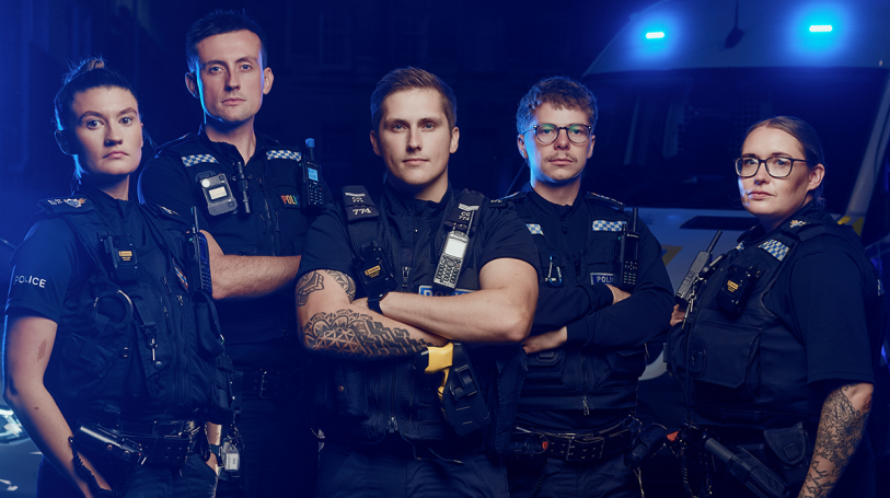 Five police officers in uniform, standing confidently with police lights in the background.