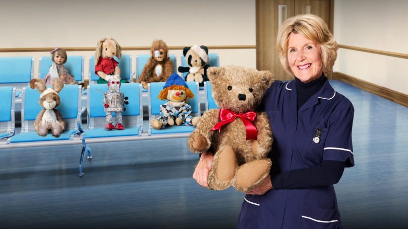 A nurse holds a teddy bear in a waiting area with various soft toys on seats.