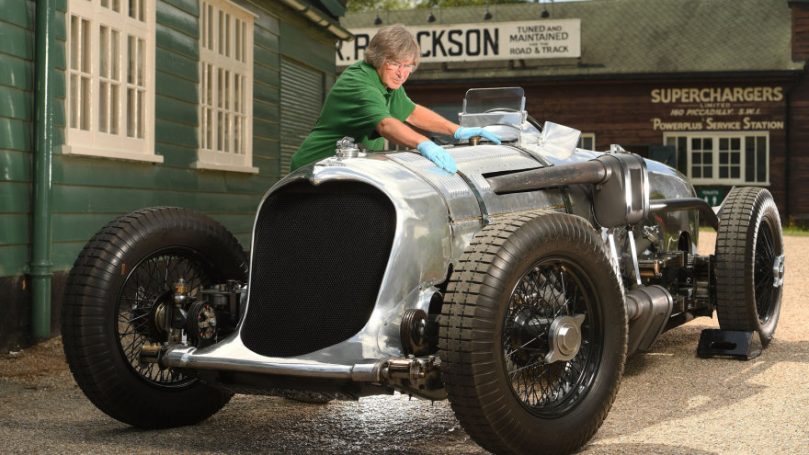 A person cleans and maintains a vintage racing car outside a workshop.