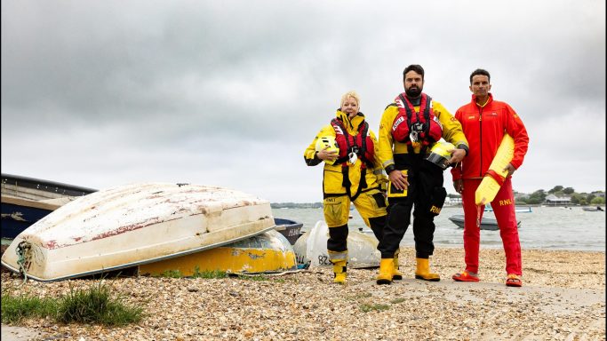 Three individuals in rescue suits stand on a beach with overturned boats in the background.