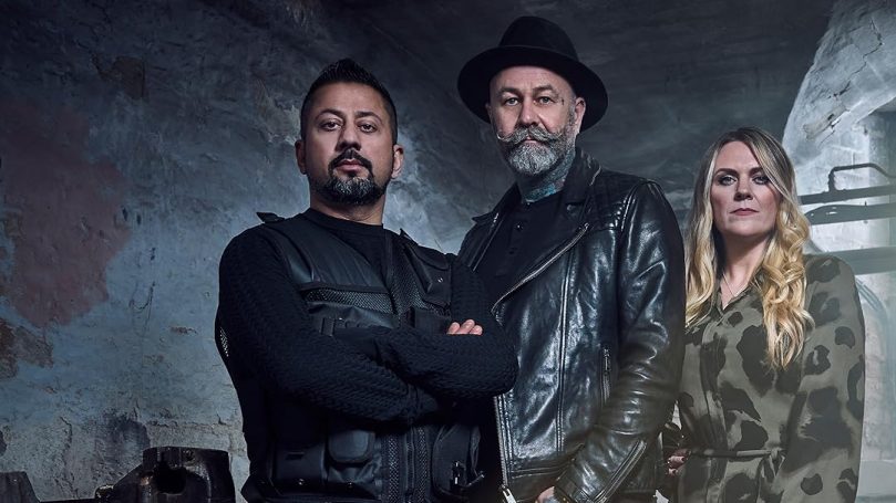 Three individuals pose together, dressed in dark clothing, against an industrial background.